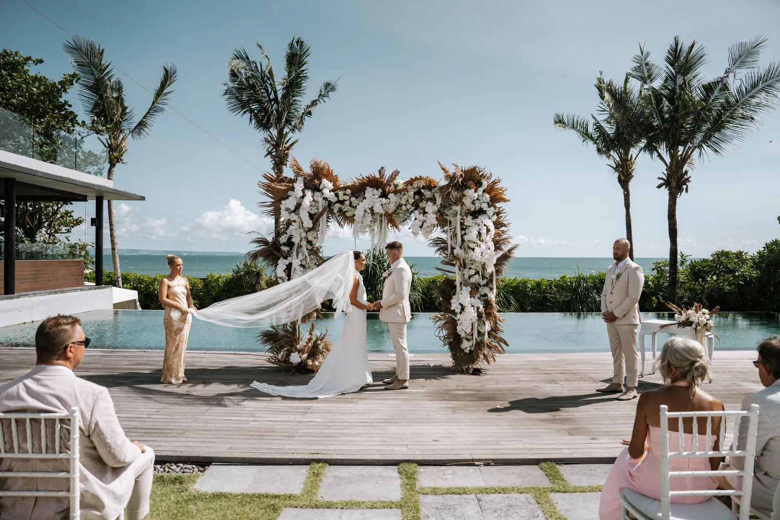 A ceremony at Arnalaya Beach House, the dried-palm arch set beside the pool with the ocean beyond