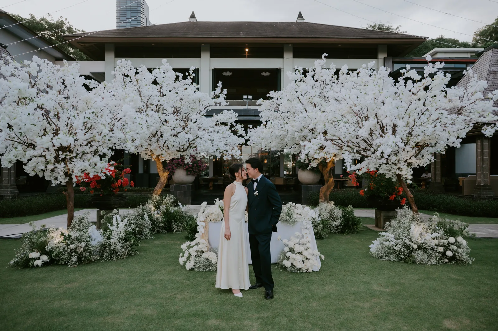 A couple together beneath blossom trees on the lawn at The Ritz-Carlton, Bali, the resort pavilion behind them